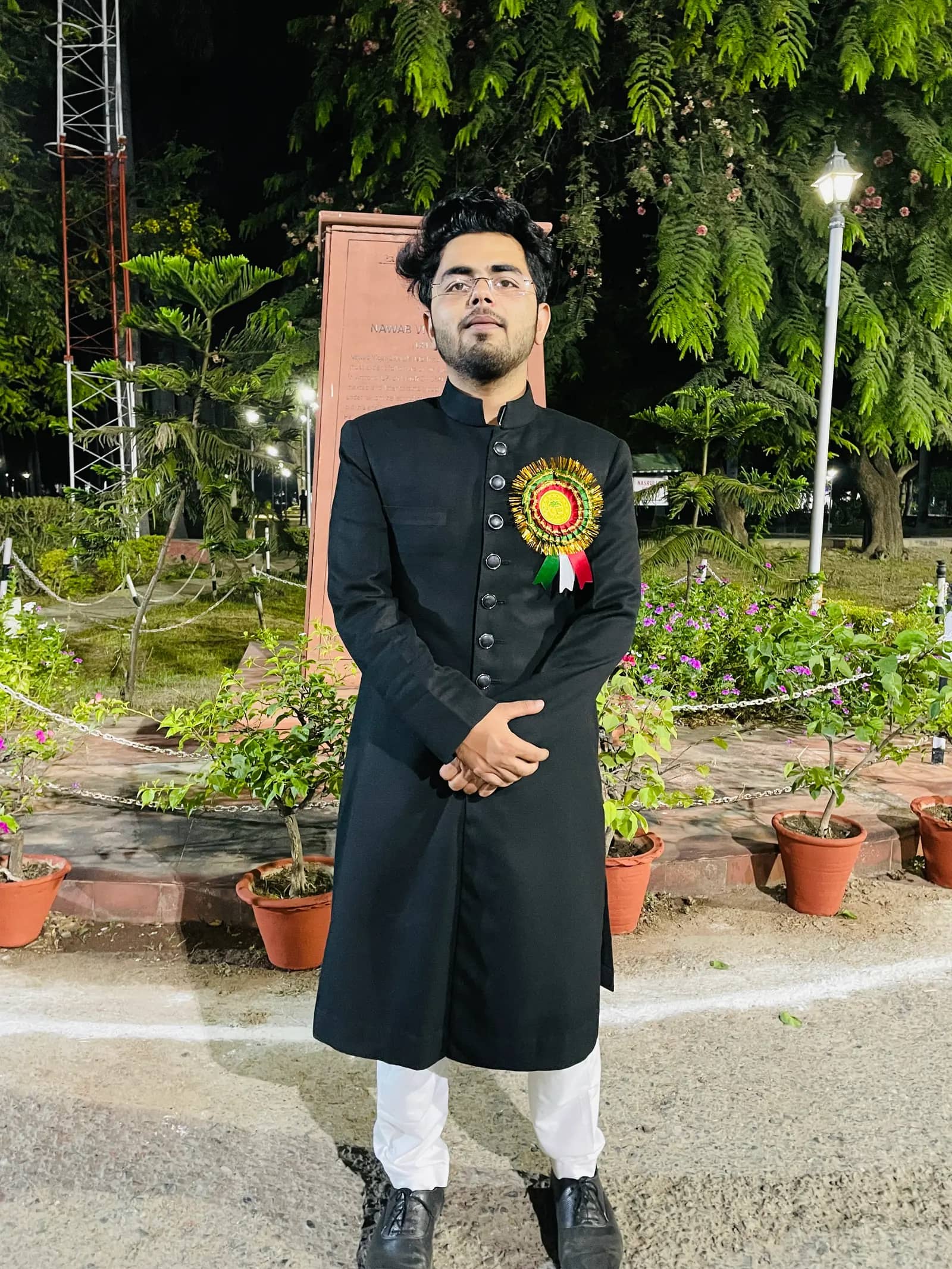 Shuja in a black sherwani with a rosette, standing in a lit garden at night.